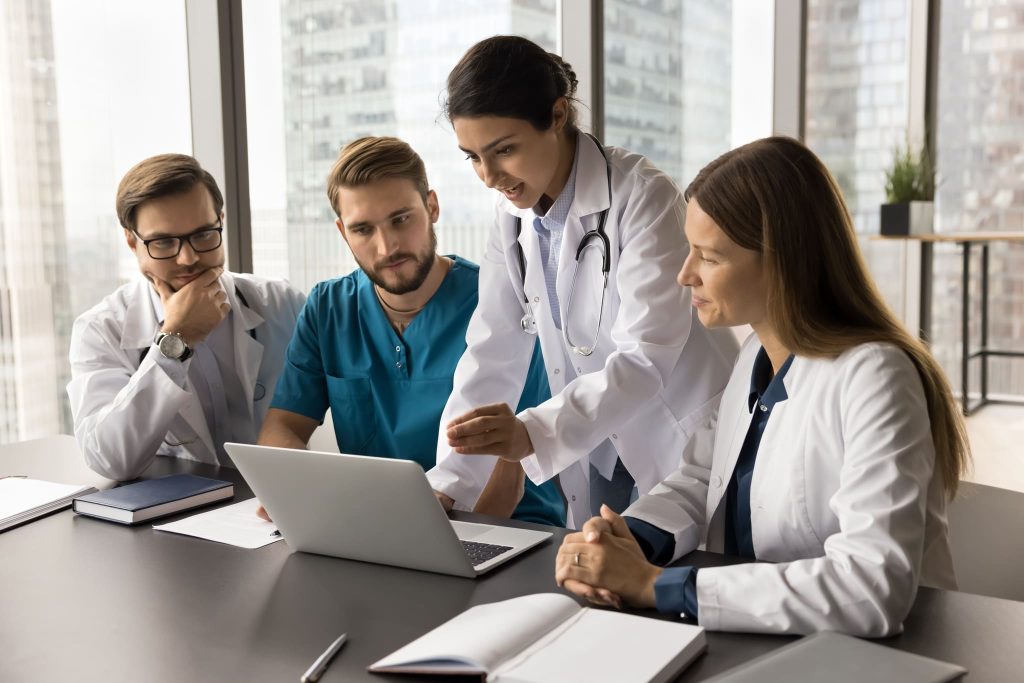 Four doctors, two men and two women, collaborate around a laptop in a brightly lit office.